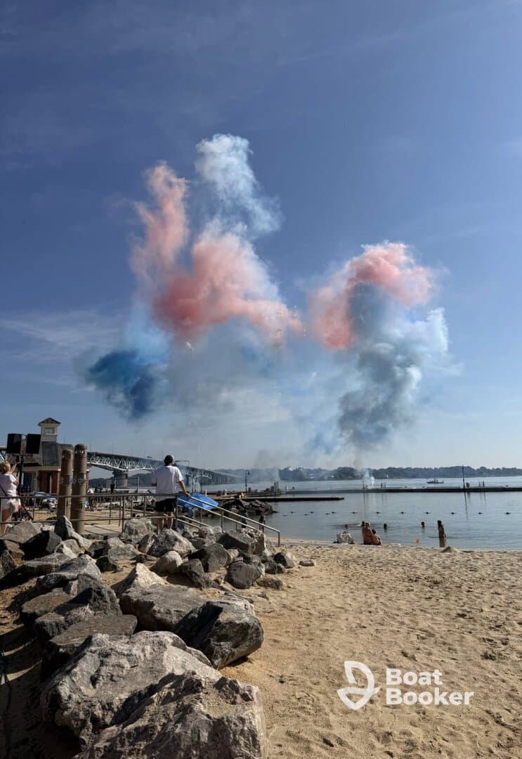 A vibrant display of colored smoke fills the sky during a beach celebration, with people enjoying the festive atmosphere by the water.
