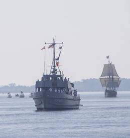 A naval vessel sails alongside a historic ship, showcasing a vibrant maritime scene with crew members visible on deck.