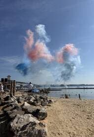 A vibrant display of colored smoke fills the sky during a beach celebration, with people enjoying the festive atmosphere by the water.