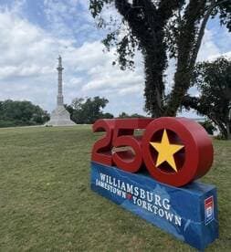A scenic view featuring a commemorative sign for the 250th anniversary of Williamsburg, Jamestown, and Yorktown, with a monument in the background.