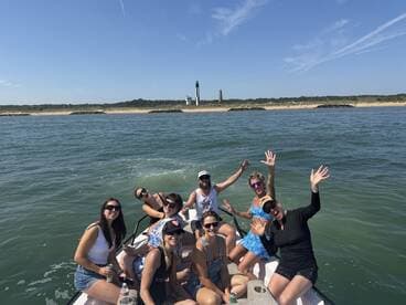 A lively group of friends enjoys a sunny day on the water, celebrating with drinks and smiles, with a picturesque lighthouse in the background.