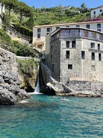 Swimmers enjoy the clear waters near a picturesque coastline featuring a charming waterfall and scenic stone architecture, perfect for a summer getaway.
