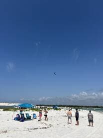 A lively group enjoys a sunny day at the beach, capturing moments while watching activities in the sky — perfect for family outings and beach fun.