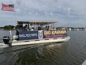 A lively tiki charter boat, 'Big Kahuna,' cruises through calm waters with guests enjoying the ride — perfect for fun outings and family adventures.