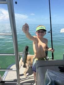 A young boy proudly holds up his catch while fishing on a boat, with a playful dog nearby — a perfect moment of joy on the water.