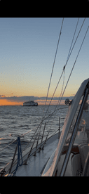 A serene view of a sailboat navigating through calm waters at sunset, with another vessel visible in the distance, creating a picturesque maritime scene.