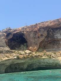 A stunning view of a rocky coastline with a cave, featuring people swimming in the crystal-clear waters — a perfect spot for exploration and adventure.