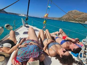 A group of friends relaxes and sunbathes on the deck of a sailboat, enjoying the beautiful turquoise waters and scenic coastline — the perfect setting for a day of sailing and relaxation.