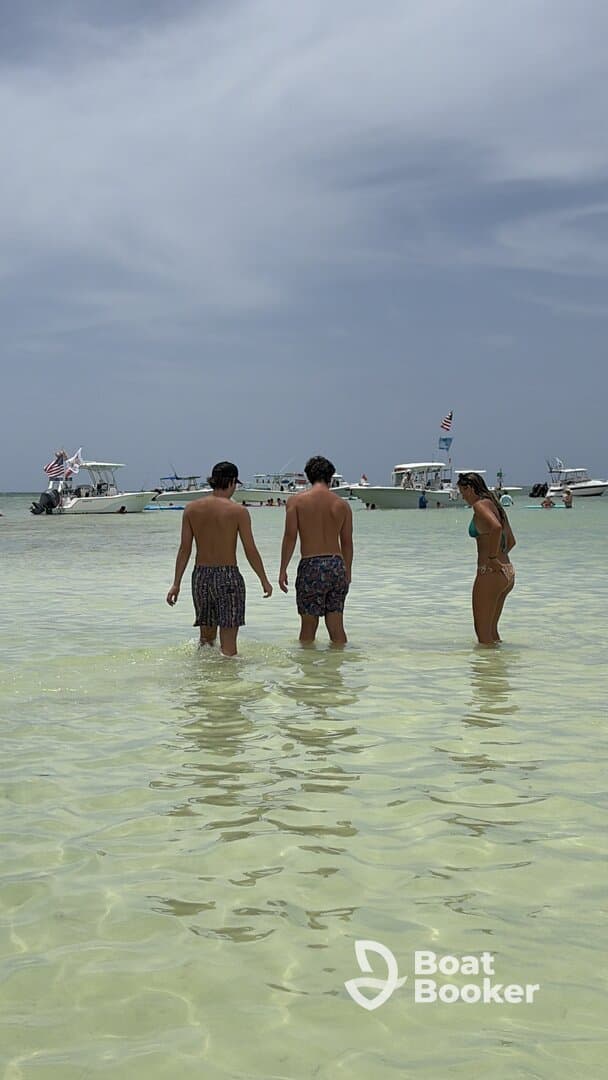 A group of friends wades in shallow water, enjoying a sunny day at the beach with boats anchored nearby — perfect for a fun day out on the water.