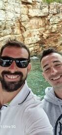 Two friends pose for a selfie on a boat with stunning rocky scenery in the background, capturing a memorable moment during their adventure.