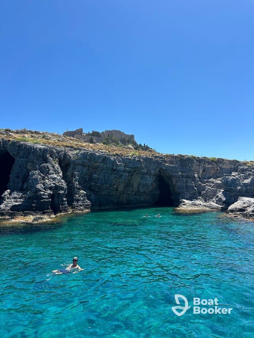 Swimmers enjoy the crystal-clear waters near stunning rock formations, with a historic castle in the background — a perfect spot for a refreshing dip and scenic views.
