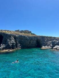 Swimmers enjoy the crystal-clear waters near stunning rock formations, with a historic castle in the background — a perfect spot for a refreshing dip and scenic views.