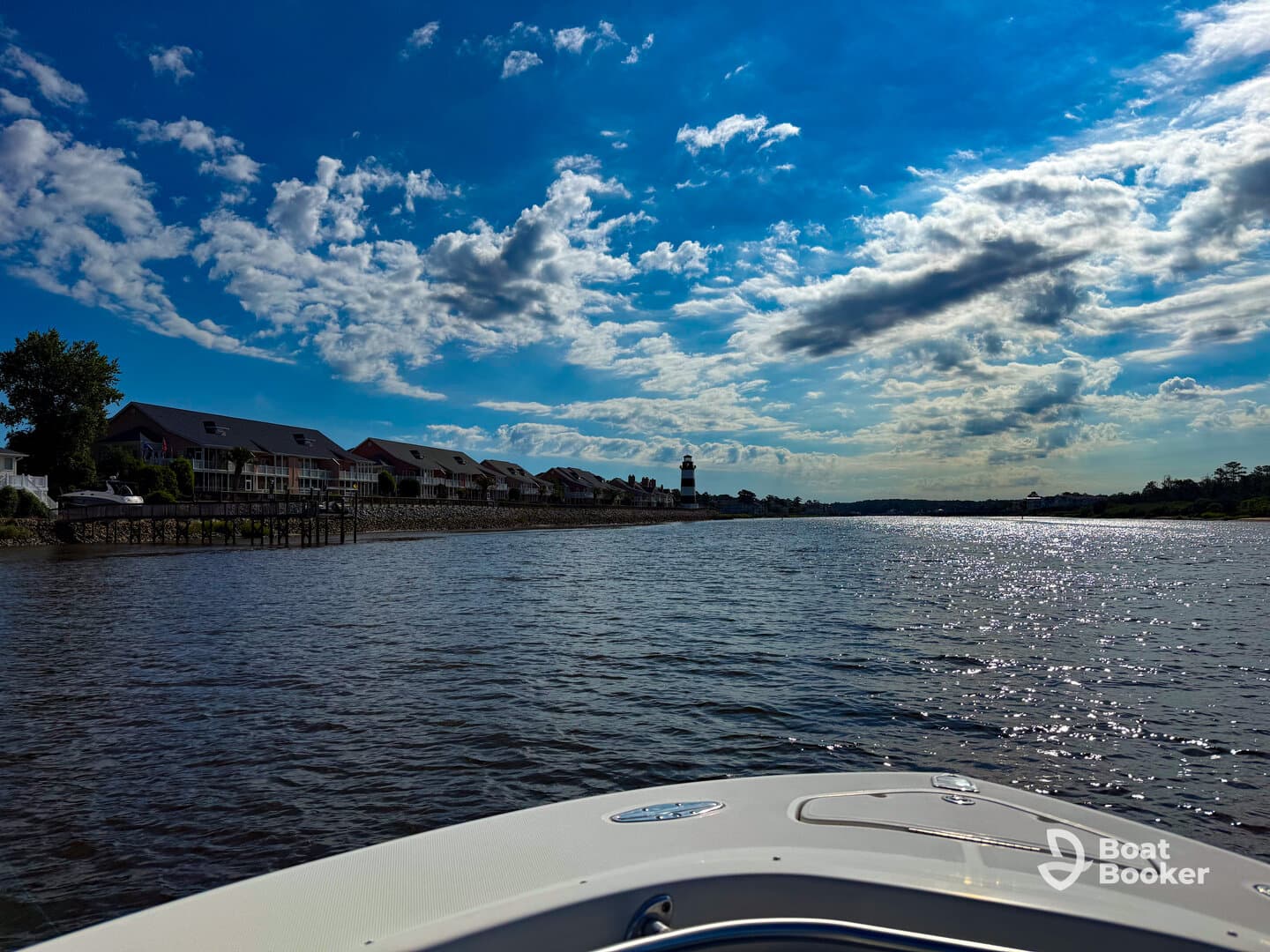 A scenic view from the bow of a boat, showcasing a tranquil waterway lined with charming homes and a lighthouse under a vibrant sky.
