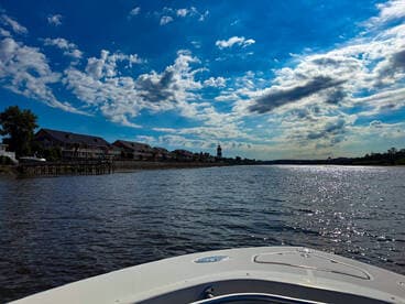 A scenic view from the bow of a boat, showcasing a tranquil waterway lined with charming homes and a lighthouse under a vibrant sky.