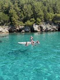 A joyful group enjoys a sunny day swimming and paddleboarding in crystal-clear waters, surrounded by lush greenery — a perfect summer getaway.