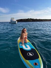 A young woman enjoys paddleboarding with a refreshing cocktail in hand, with a boat anchored in the background — a perfect day on the water.