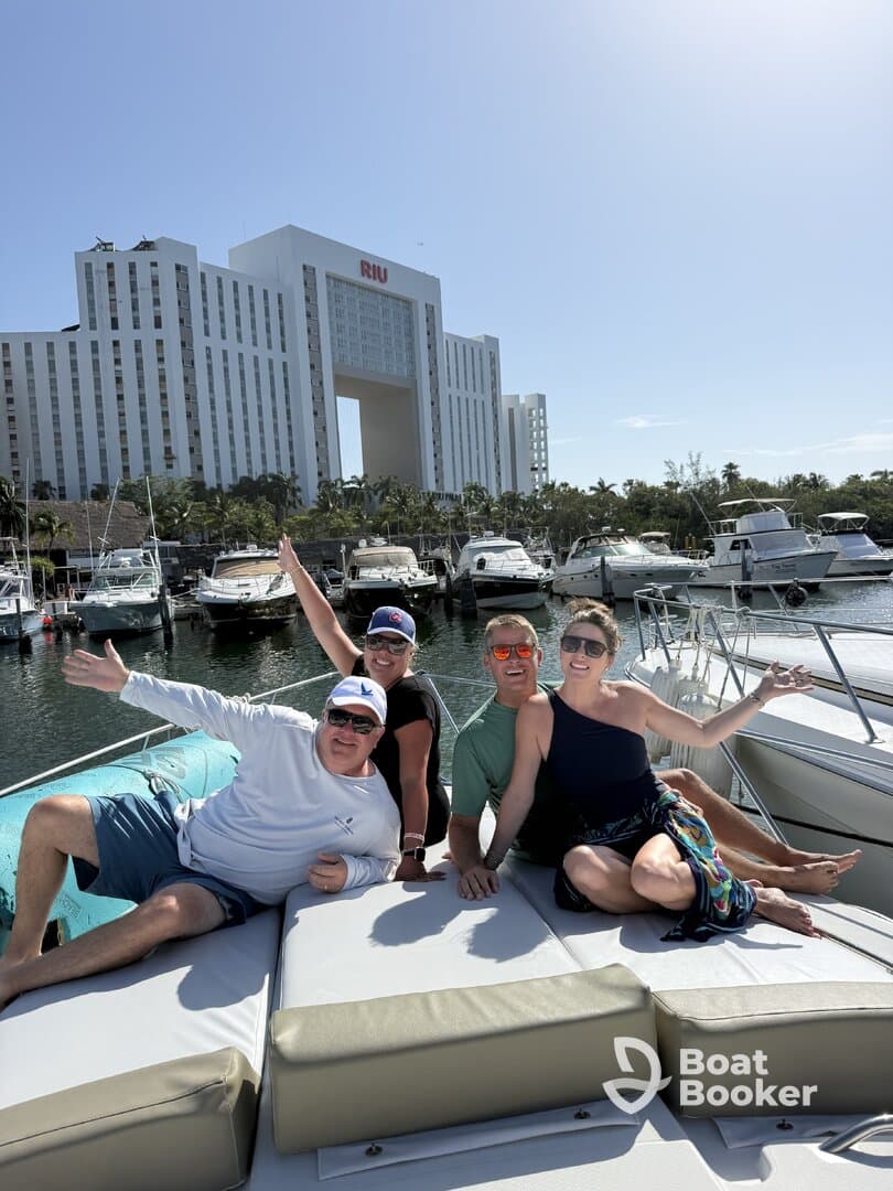 A cheerful group of friends enjoys their time aboard a boat in a marina, celebrating with smiles and waves against a backdrop of luxury yachts and a hotel.