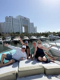 A cheerful group of friends enjoys their time aboard a boat in a marina, celebrating with smiles and waves against a backdrop of luxury yachts and a hotel.