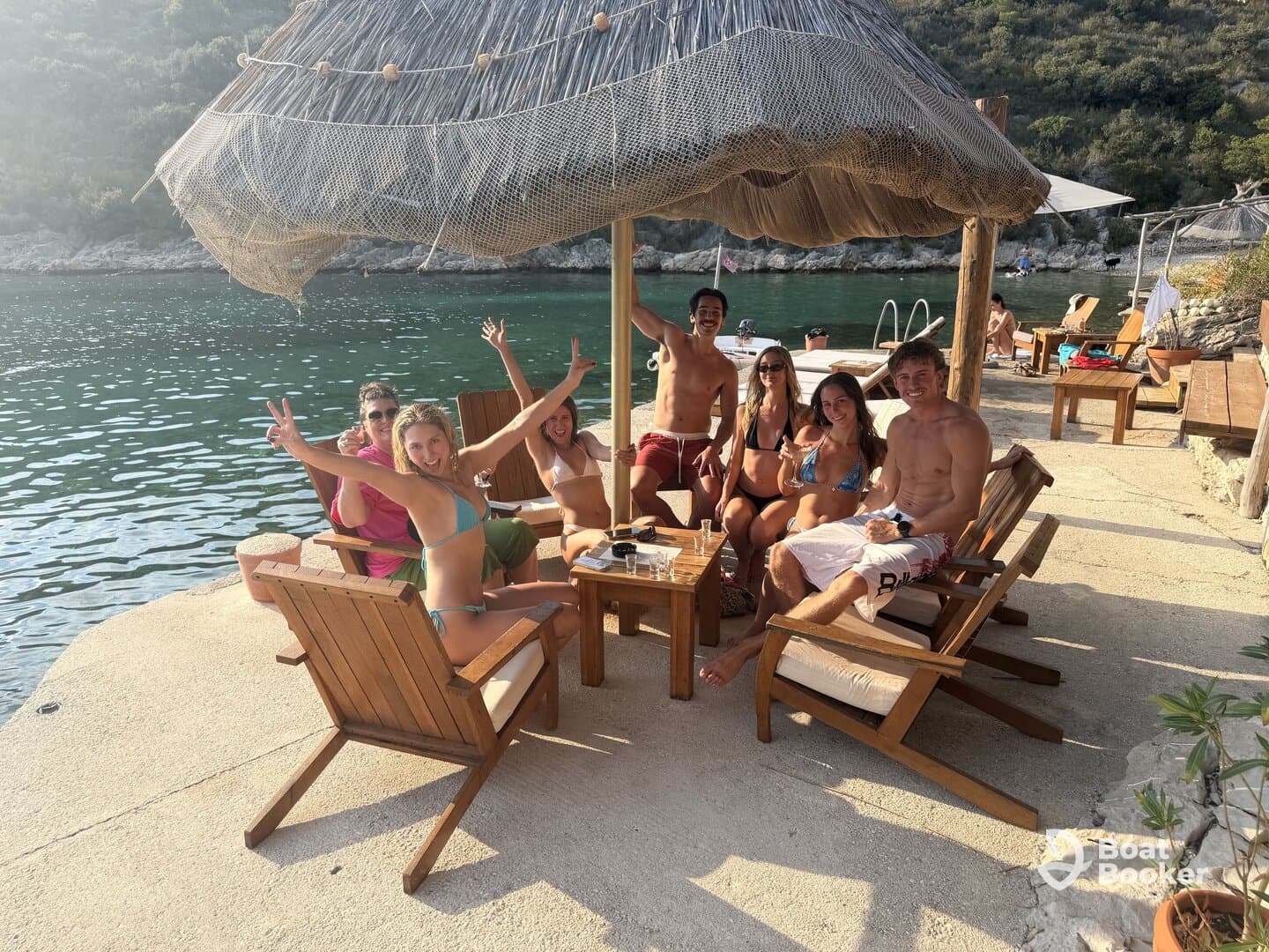 A lively group enjoys drinks and sunshine by the water, celebrating together in a picturesque beach setting — perfect for summer gatherings and fun outings.