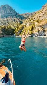 A woman joyfully leaps into the crystal-clear waters from a boat, surrounded by stunning cliffs and lush greenery — a perfect moment of adventure on the water.