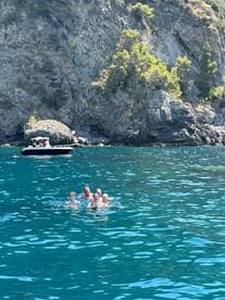 A joyful group swims in the crystal-clear waters near a rocky coastline, with a boat anchored nearby — perfect for family adventures and summer fun.