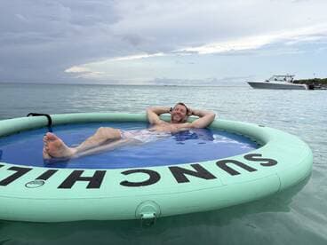 A man relaxes on a floating mat in the ocean, enjoying the serene waters with a boat anchored nearby — a perfect scene for a day of leisure on the water.
