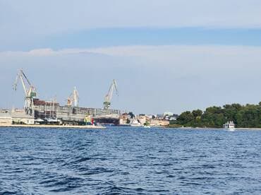 A scenic view of a harbor featuring a boat near the shore, with shipbuilding cranes and a coastal town in the background, showcasing the maritime industry.