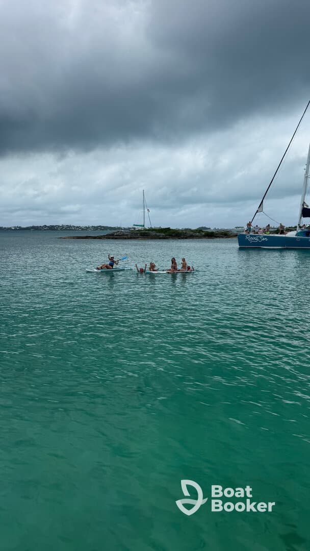 A lively group enjoys paddleboarding in clear turquoise waters, with a sailboat in the background, creating a perfect setting for water activities and fun in the sun.