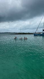 A lively group enjoys paddleboarding in clear turquoise waters, with a sailboat in the background, creating a perfect setting for water activities and fun in the sun.