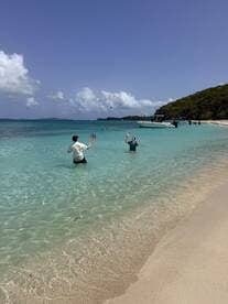 Two friends enjoy a playful day at the beach, tossing a ball in the shallow waters while a boat is anchored nearby — a perfect scene for summer fun and relaxation.