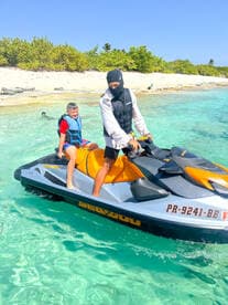 A child enjoys a thrilling ride on a jet ski with an adult, surrounded by clear turquoise waters and a sandy beach — perfect for family adventures on the water.