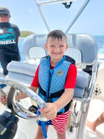 A young boy joyfully steers a boat while wearing a life jacket, capturing the excitement of family boating adventures.