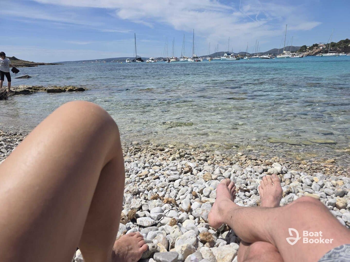 A couple relaxes on a rocky beach, enjoying the serene view of boats anchored in the turquoise waters — a perfect getaway for sun and sea lovers.