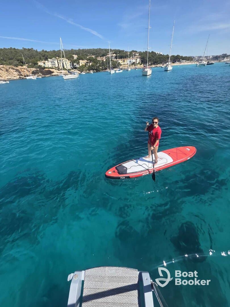 A paddleboarder enjoys the crystal-clear waters, surrounded by scenic views of the coastline and anchored boats — a perfect snapshot of a fun day on the water.