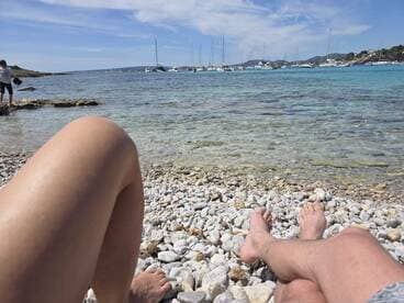 A couple relaxes on a rocky beach, enjoying the serene view of boats anchored in the turquoise waters — a perfect getaway for sun and sea lovers.