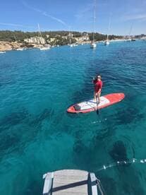 A paddleboarder enjoys the crystal-clear waters, surrounded by scenic views of the coastline and anchored boats — a perfect snapshot of a fun day on the water.