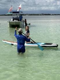 A child enjoys paddleboarding while an adult assists in shallow waters, creating a memorable experience on the water with friends and family.