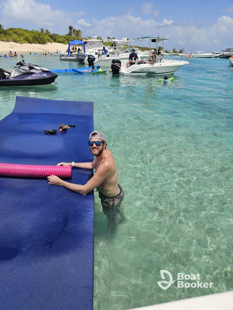 A man enjoys a sunny day at the beach, interacting with a floating mat while boats and fellow beachgoers create a lively atmosphere — perfect for summer fun and water activities.
