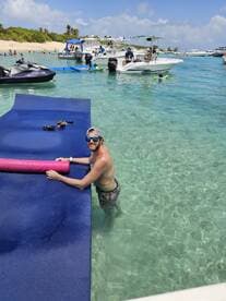 A man enjoys a sunny day at the beach, interacting with a floating mat while boats and fellow beachgoers create a lively atmosphere — perfect for summer fun and water activities.