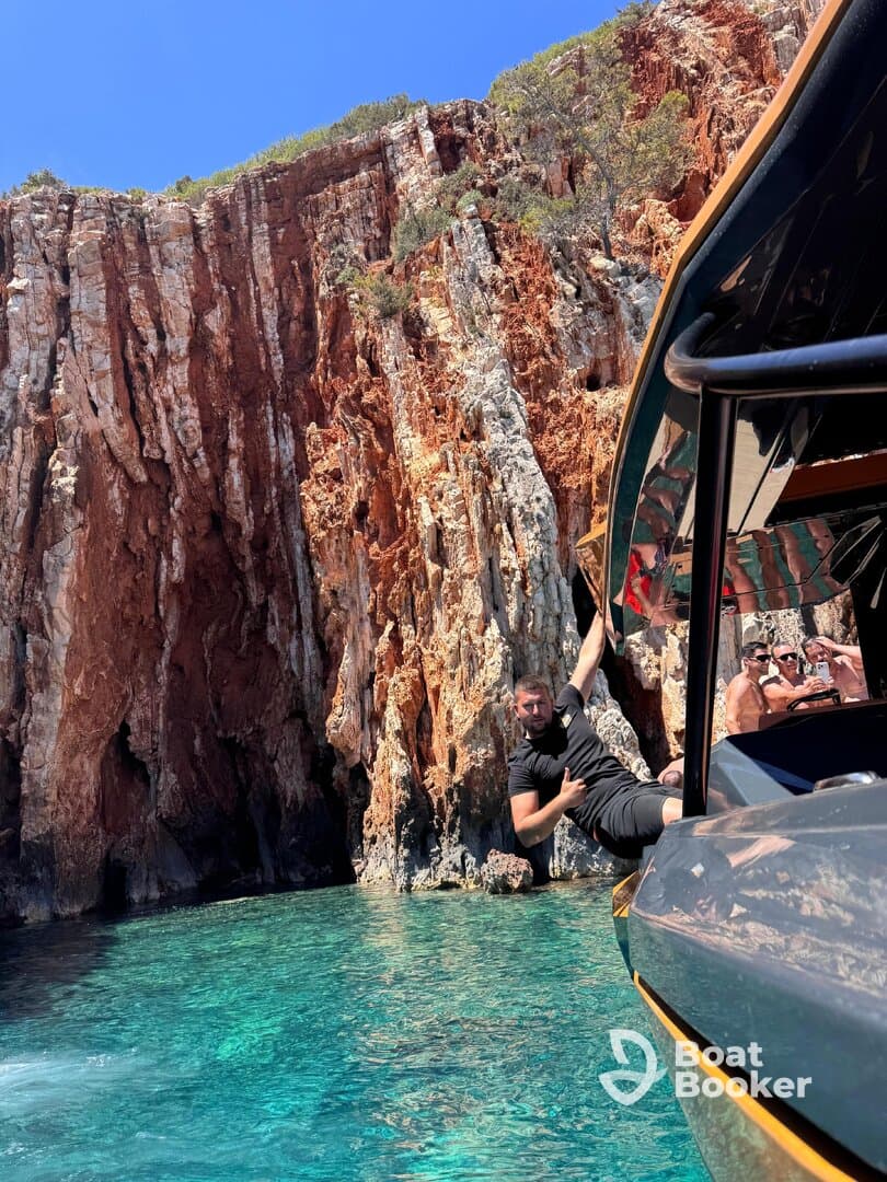 A man enjoys a thrilling moment hanging off the side of a boat near stunning cliffs, with clear turquoise waters creating a perfect backdrop for adventure.