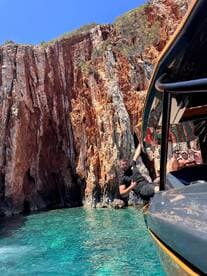 A man enjoys a thrilling moment hanging off the side of a boat near stunning cliffs, with clear turquoise waters creating a perfect backdrop for adventure.