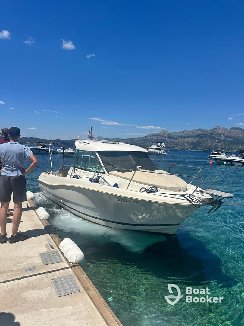 A boat approaches the dock with two people observing the arrival, set against a stunning backdrop of clear blue waters and mountains.