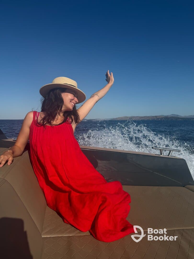 A joyful woman in a red dress enjoys a sunny day on a boat, capturing the moment with her phone against a stunning ocean backdrop.