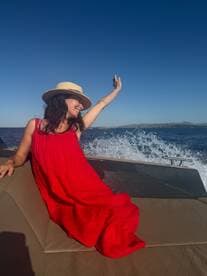 A joyful woman in a red dress enjoys a sunny day on a boat, capturing the moment with her phone against a stunning ocean backdrop.