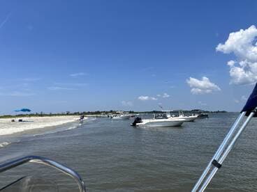 A vibrant scene at a sandbar with multiple boats anchored nearby, where people enjoy the beach and water activities under a clear blue sky.