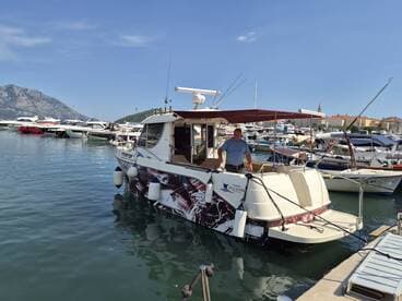 A fishing boat named 'Predator' docked in a marina, with a crew member preparing for the day. The scenic backdrop features mountains and other vessels, ideal for fishing adventures.