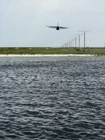 A distant view of an aircraft approaching for landing over a body of water, with power lines and vehicles visible in the background.