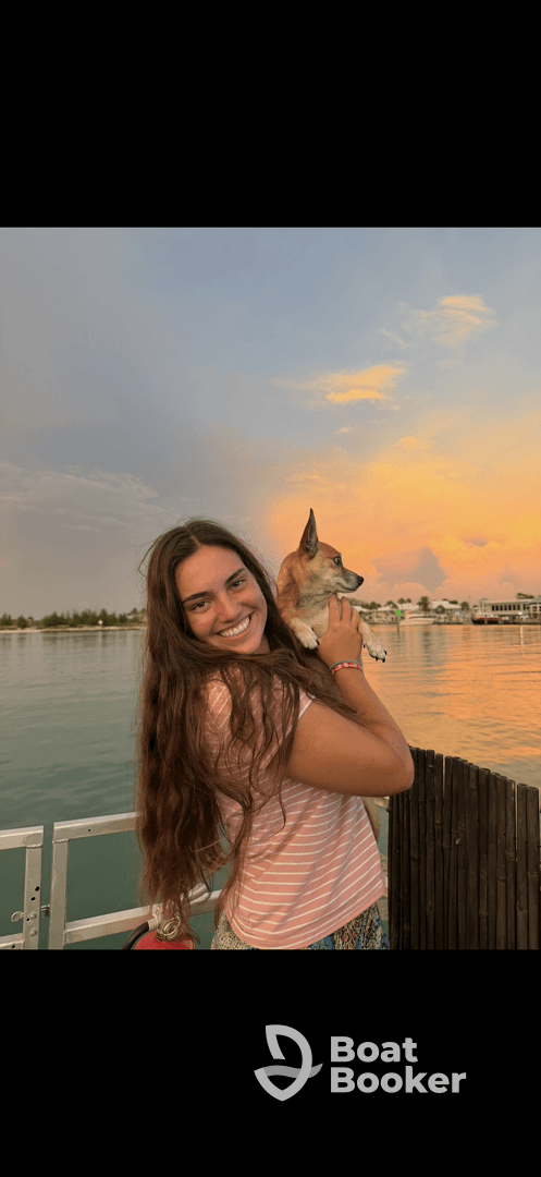 A young woman smiles while holding her dog against a beautiful sunset backdrop, capturing a heartwarming moment of companionship by the water.