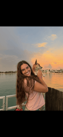 A young woman smiles while holding her dog against a beautiful sunset backdrop, capturing a heartwarming moment of companionship by the water.