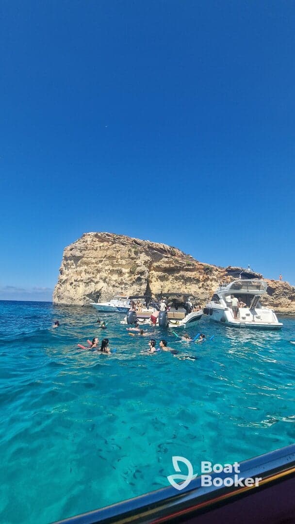 A lively scene of guests enjoying a swim in crystal-clear waters near a rocky coastline, with boats anchored nearby — perfect for snorkeling and beach fun.